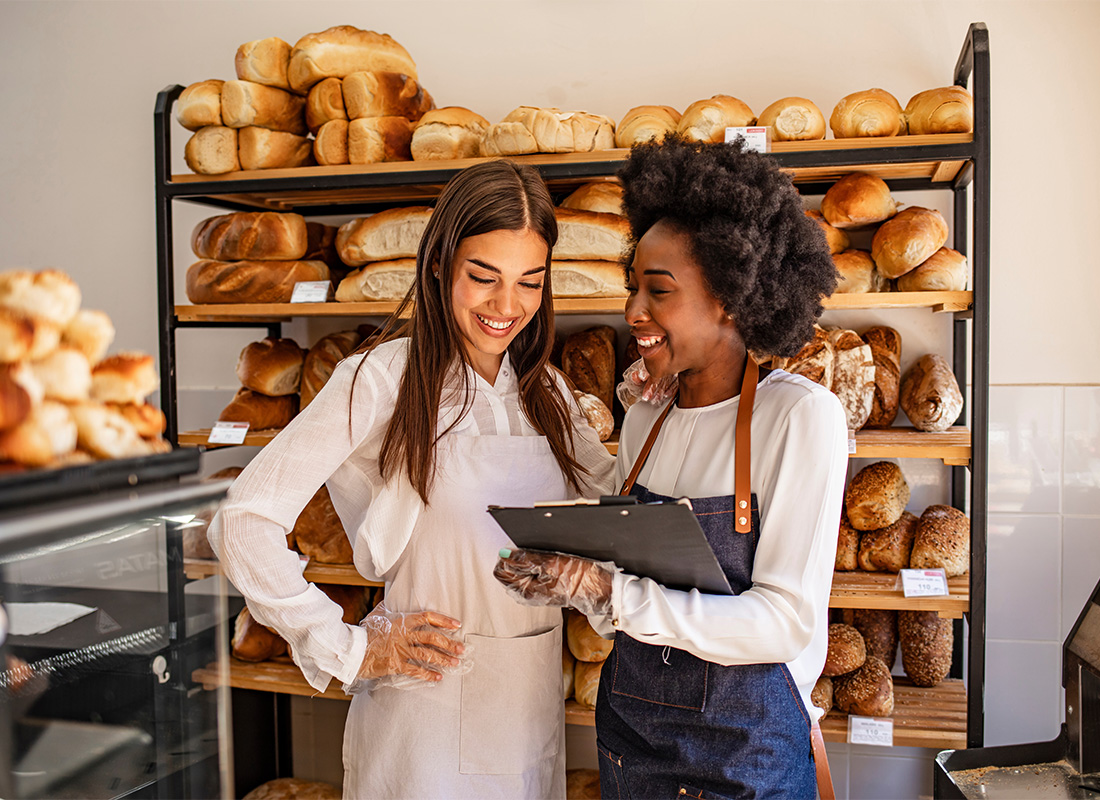 Business Insurance - Portrait of Two Smiling Young Women Standing Inside a Bakery Next to Loaves of Bread While Holding a Tablet